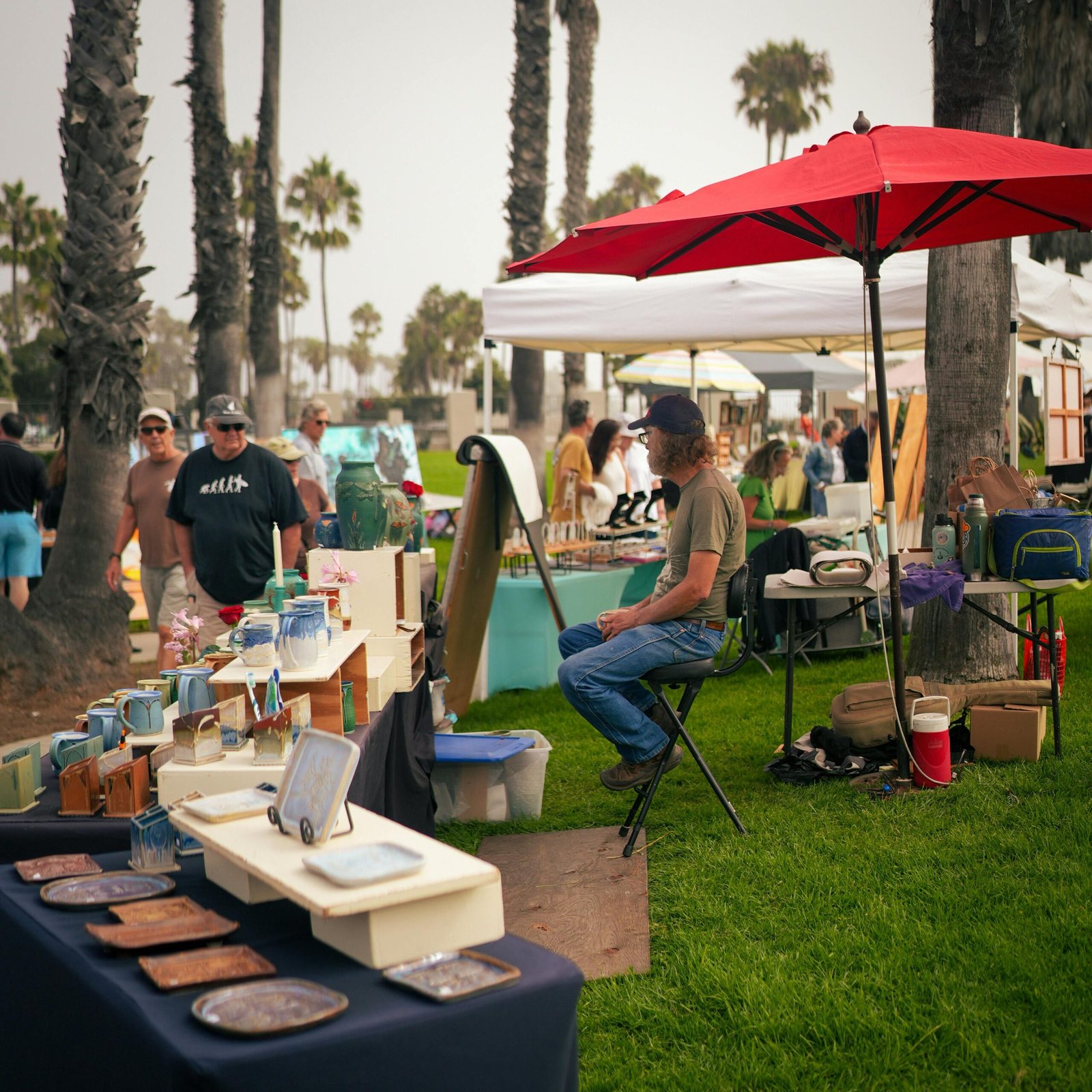 Vibrant outdoor market scene with art stalls and shoppers under palm trees.